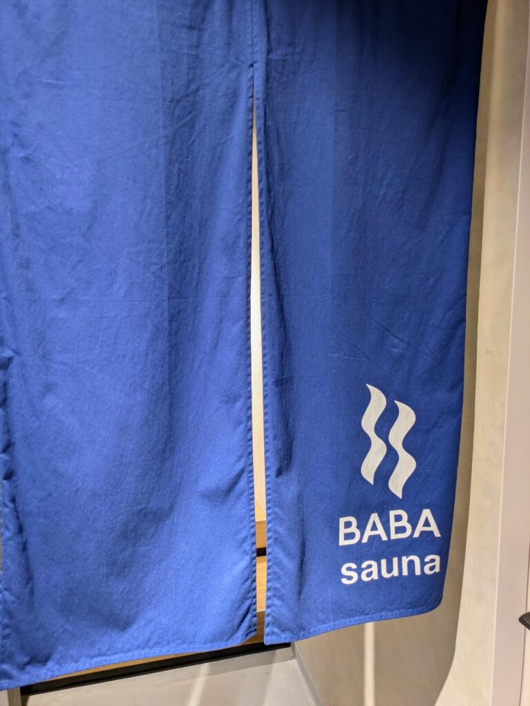 馬場サウナの入り口にかかっている、青い暖簾。暖簾にはBABA saunaのロゴと文字が白で描かれている。