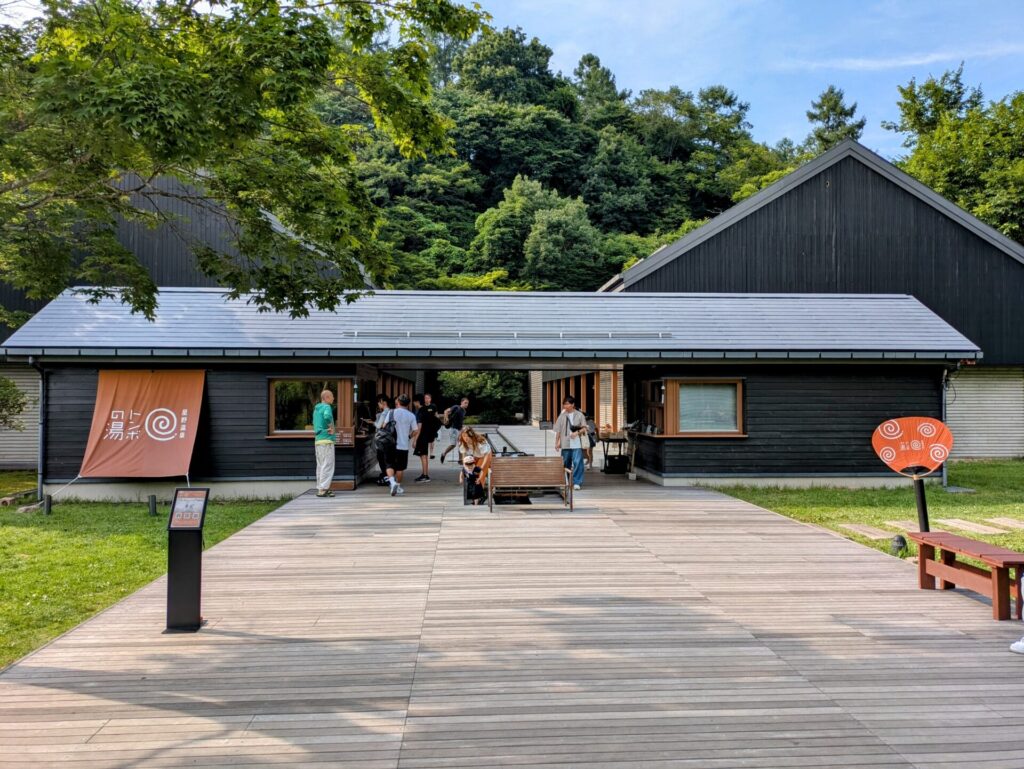 長野県軽井沢にある星野温泉トンボの湯の入り口。黒い木造建築と木製のデッキ、そして緑豊かな自然が調和したモダンな外観。