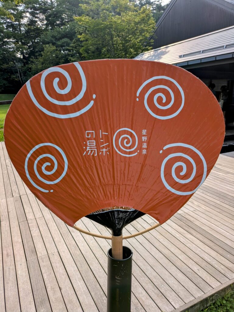 星野温泉トンボの湯のロゴが描かれた団扇型の看板。オレンジ色の団扇に渦巻き模様と「トンボの湯」「星野温泉」の文字がデザインされている。