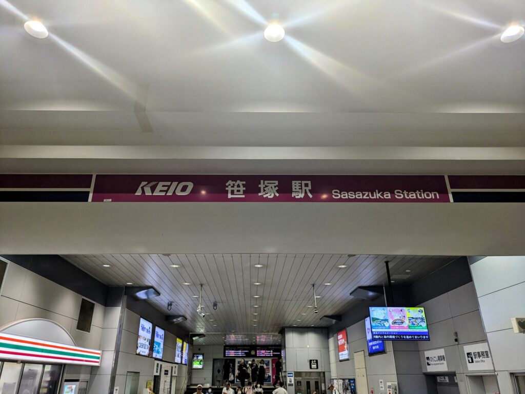 京王線笹塚駅構内の様子。紫色の看板に「KEIO 笹塚駅 Sasazuka Station」と日本語と英語で表示されている。天井には白い照明が複数あり、下には駅の改札や店舗が見える。多くの人が行き来している様子が伺える。