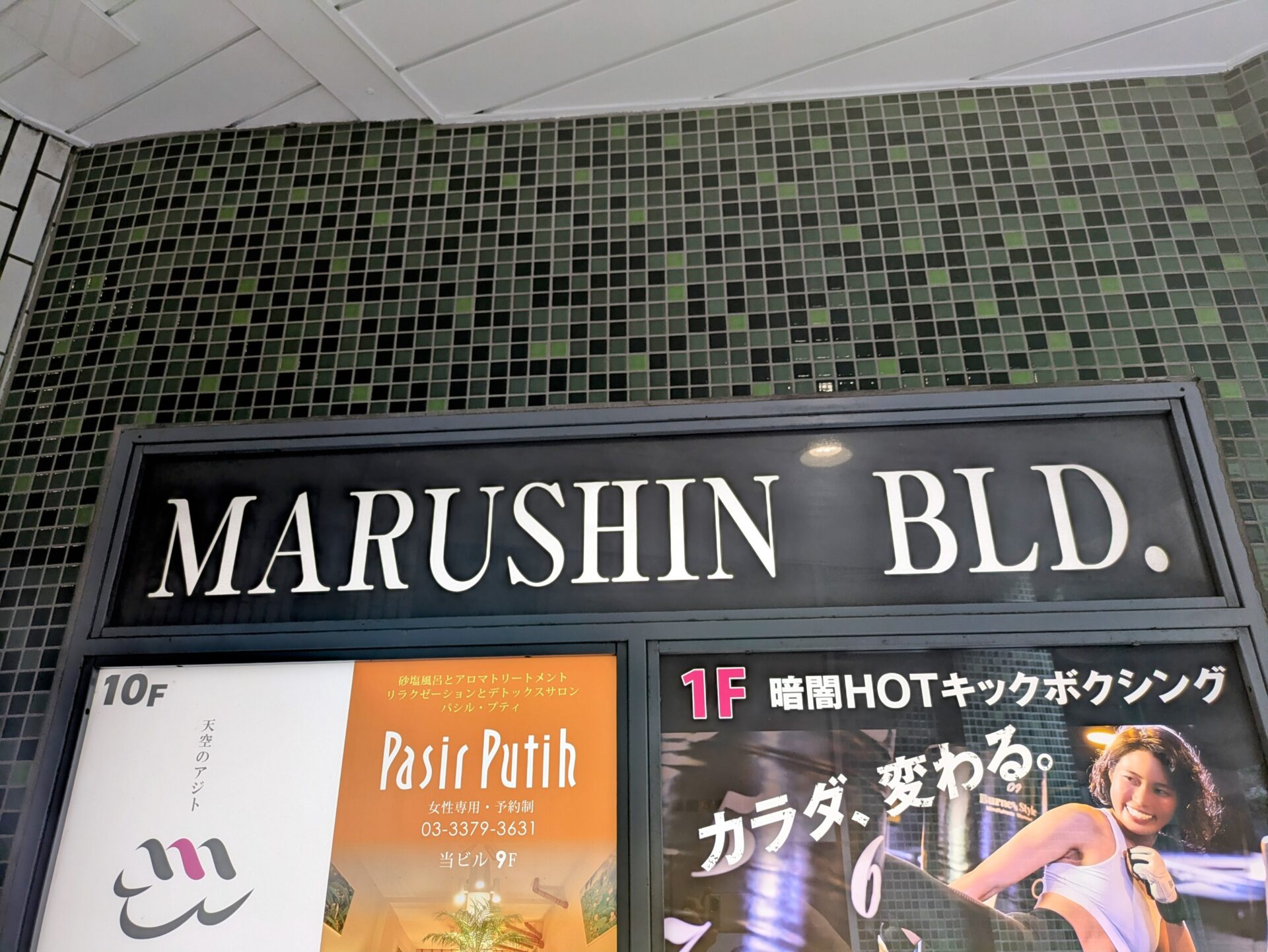 「マルシンビル」の看板が写っている。上部には「MARUSHIN BLD.」と書かれており、その下にはテナント情報が掲示されている。左側の「10F」には「天空のアジト マルシンスパ」のロゴと文字、中央には9Fの「Pasir Putih」、右側には1Fの「暗闇HOTキックボクシング」の看板が見える。
