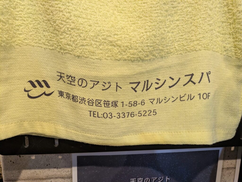 「天空のアジト マルシンスパ」のロゴと住所(東京都渋谷区笹塚1-58-6 マルシンビル10F)、電話番号(TEL: 03-3376-5225)が印字された黄色のタオル。