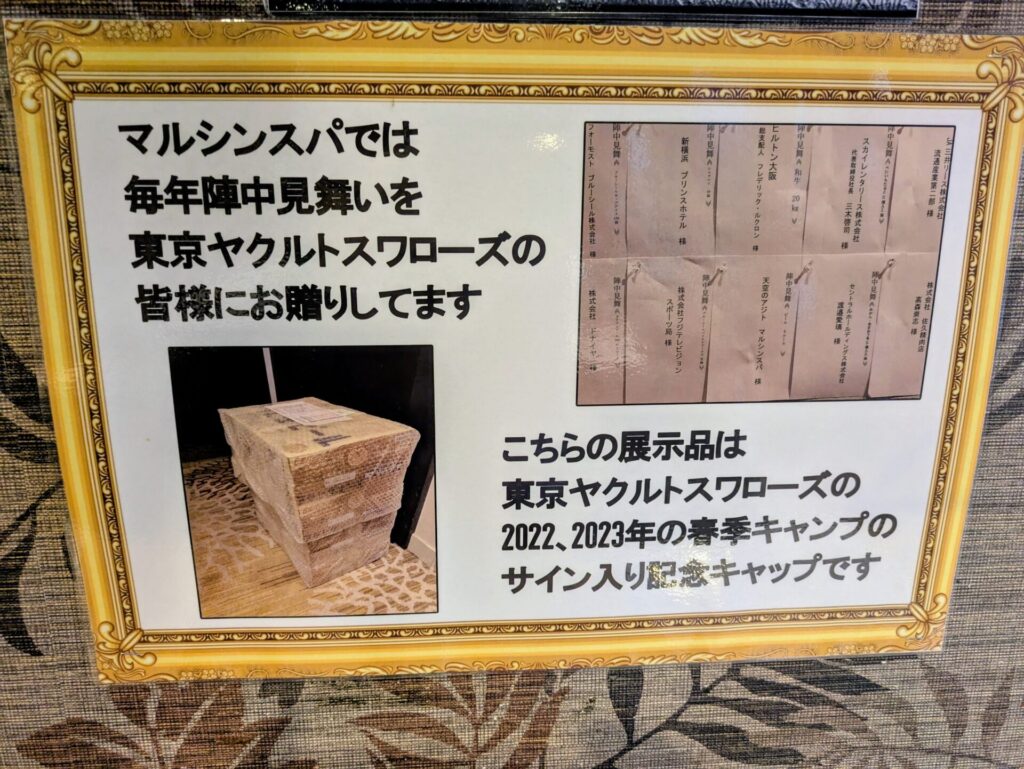 「マルシンスパでは毎年陣中見舞いを東京ヤクルトスワローズの皆様にお贈りしてます」と書かれたボード。その下には、積み重ねられた記念キャップと、「東京ヤクルトスワローズの2022,2023年の春季キャンプのサイン入り記念キャップです」という説明文が添えられている。右側には、キャップが入っていたと思われる袋が複数写っている。