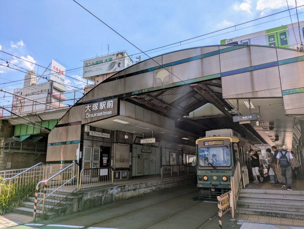 東京都豊島区、JR大塚駅前の都電荒川線「大塚駅前」停留所。レトロな雰囲気の電車が停車しており、乗降客がホームにいる。上空にはJR線の高架橋が見え、電車の色や駅名の看板が鮮明に写っている。