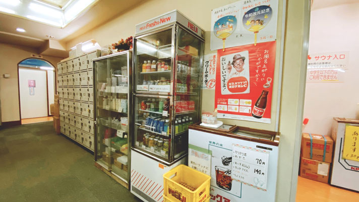 昭和レトロな雰囲気の日本の銭湯にある休憩スペース。ドリンクやお風呂用品を販売している古いショーケースと、たくさんのロッカーが並んでいる。壁には懐かしいポスターが貼られ、入浴後にくつろぐ様子が想像できる。
