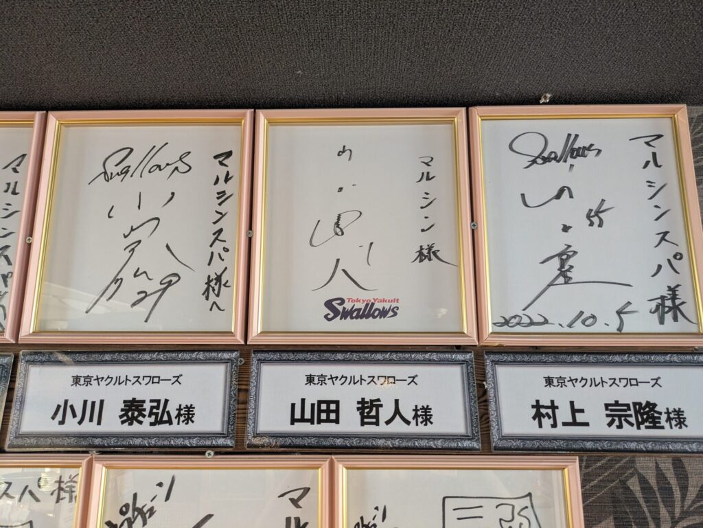 東京ヤクルトスワローズの選手たちのサイン色紙が額縁に飾られている。左から、小川泰弘投手、山田哲人選手、村上宗隆選手のサインと、それぞれ「マルシンスパ様へ」と書かれたメッセージが見える。特に村上宗隆選手のサインには、2022年10月5日のサインが見える。