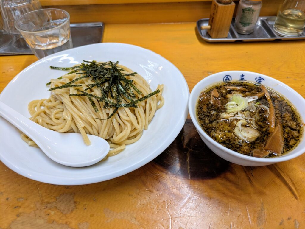 東京都豊島区、JR大塚駅近くにある人気ラーメン店「喜富」のつけ麺。ツヤのある太麺の上に刻み海苔がトッピングされ、横には具材がたっぷりの濃厚な魚介豚骨スープが添えられている。食欲をそそる美味しそうな写真。